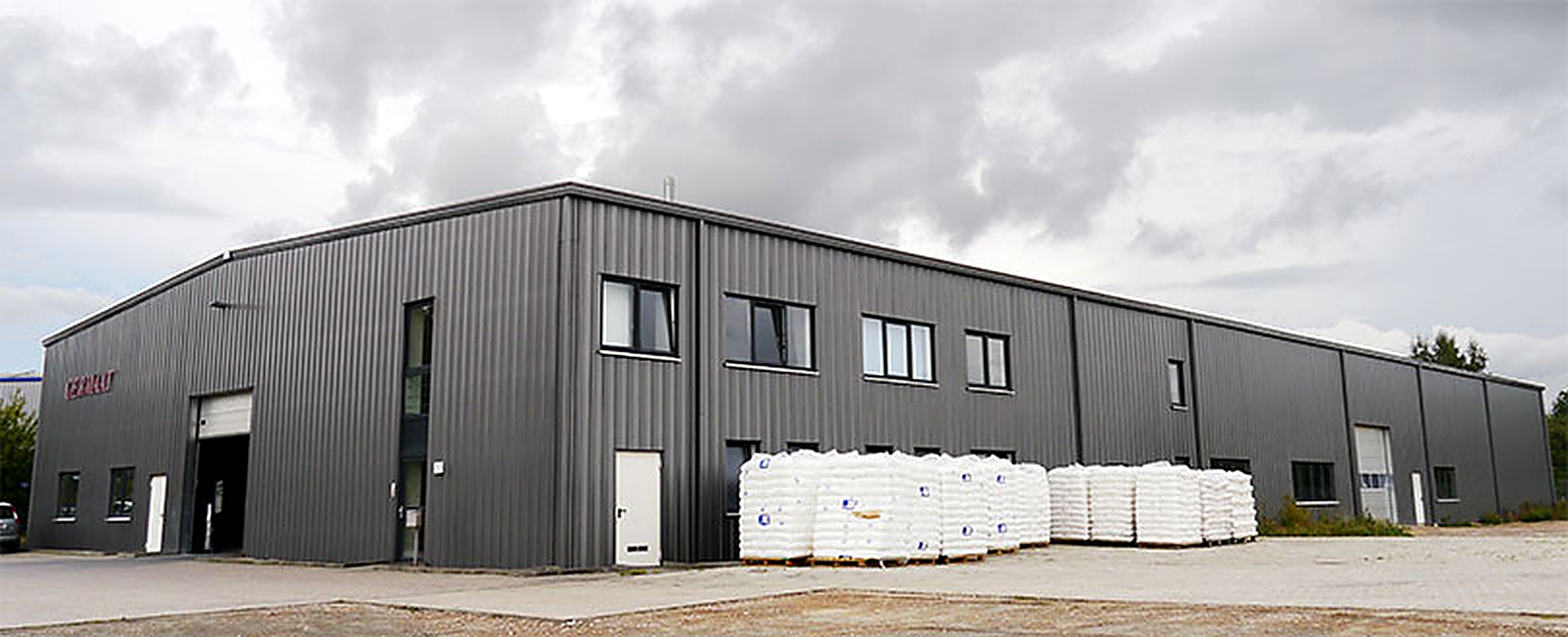 Die dunkelgraue Produktionshalle von POLYNOVA Compounds in der Robert-Bunsen-Straße in Schwerin, davor palettierte Säckeware.